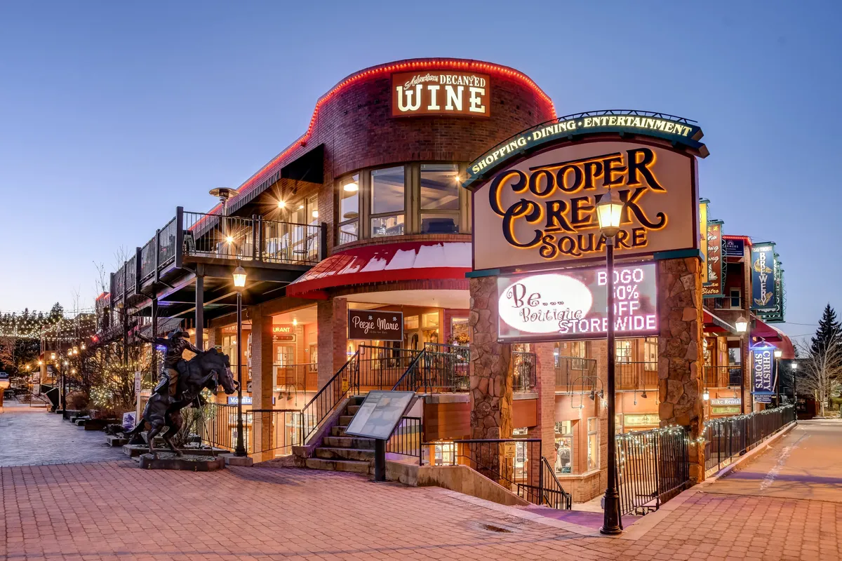 A lively downtown scene featuring the Cooper Creek Square sign, a wine shop, public sculpture, and bustling pedestrian activity. The area is welcoming and animated, ideal for shopping, dining, and community entertainment.