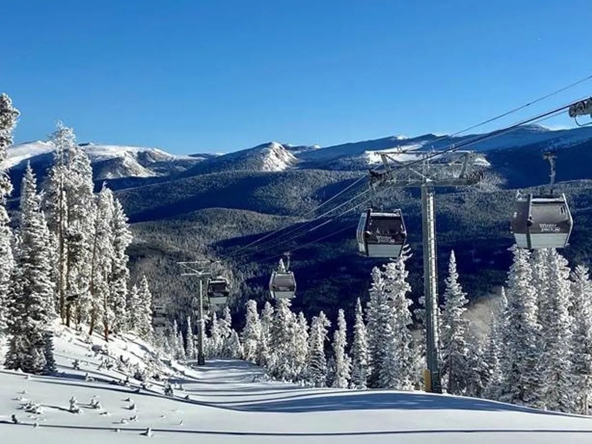 A snow-covered landscape featuring a ski lift ascending the mountain, flanked by snowy trees and distant peaks under a clear sky. The image captures the essence of a bustling winter resort with skiing and snowboarding opportunities right out the door.