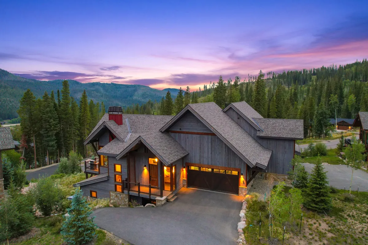 The image showcases the elegant facade of a luxury single-family villa nestled among tall, serene trees, with a spacious driveway leading up to the home. The setting sun bathes the property in a warm, golden glow, highlighting the inviting porch—perfect for relaxing or gathering with friends and family while surrounded by nature.