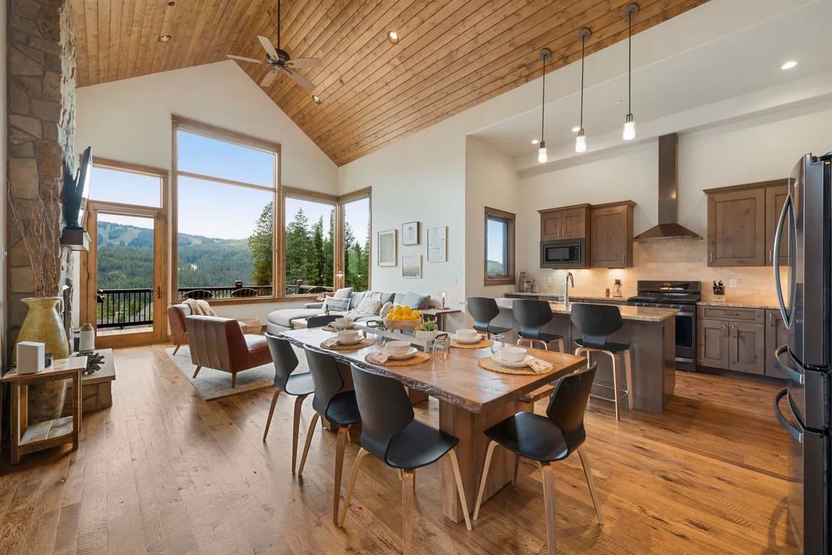 A dining area set for a meal, positioned by large windows offering panoramic mountain views. The wooden beams and modern kitchen appliances in the background unite the kitchen and dining elements for a seamless entertaining experience.