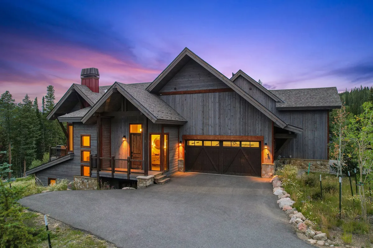 A cozy exterior shot of the home with a wooden facade, large windows, a garage door, and a front porch set against a natural backdrop. The image evokes a sense of arrival and adventure, hinting at nearby trails and the inviting wilderness beyond.