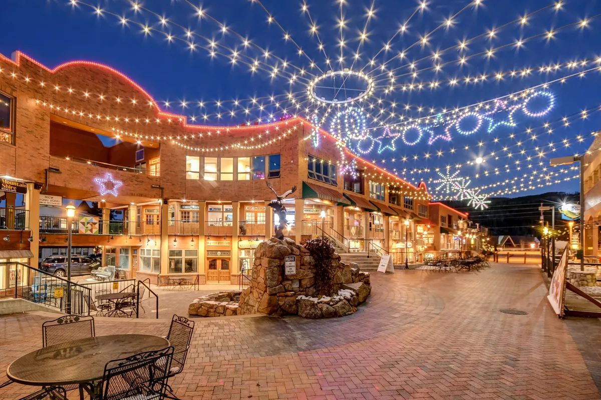 A lively, festive outdoor plaza with string lights overhead, decorative star shapes, a stone fountain, and a brick pathway. The area is set for social gatherings, seasonal events, and community enjoyment, reflecting the vibrancy of downtown Winter Park and its events during summer and shoulder seasons.