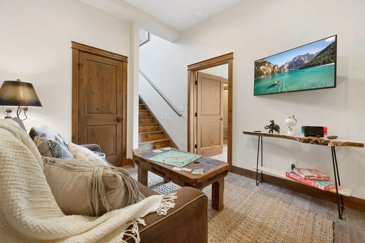 A warm, welcoming living area with a large sofa adorned with decorative pillows, a wooden coffee table set up for games or snacks, a television displaying a scenic view, and a staircase leading to the upper level. The space is bathed in natural light from the large windows.