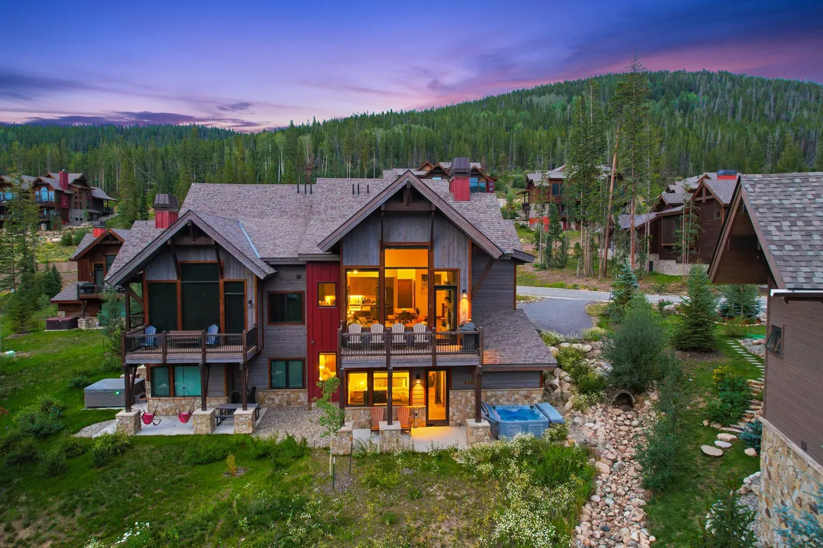 An expansive exterior view of the home featuring large windows, a welcoming outdoor seating area, a bubbling hot tub, and a landscaped garden. The mountains are visible in the background, and the space is bathed in soft, natural light, promising both relaxation and social opportunities.