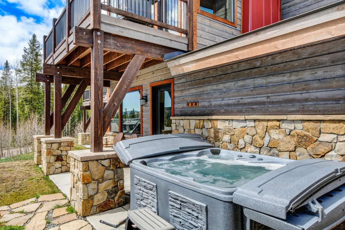 This image features a serene exterior scene with a private hot tub set on a wooden deck, bordered by a stone wall. The setting looks out onto nature, with the peacefulness of the surrounding landscape visible. The scene captures the inviting glow of the hot tub, suggesting relaxation in all weather, with potential for mountain or starlit views.