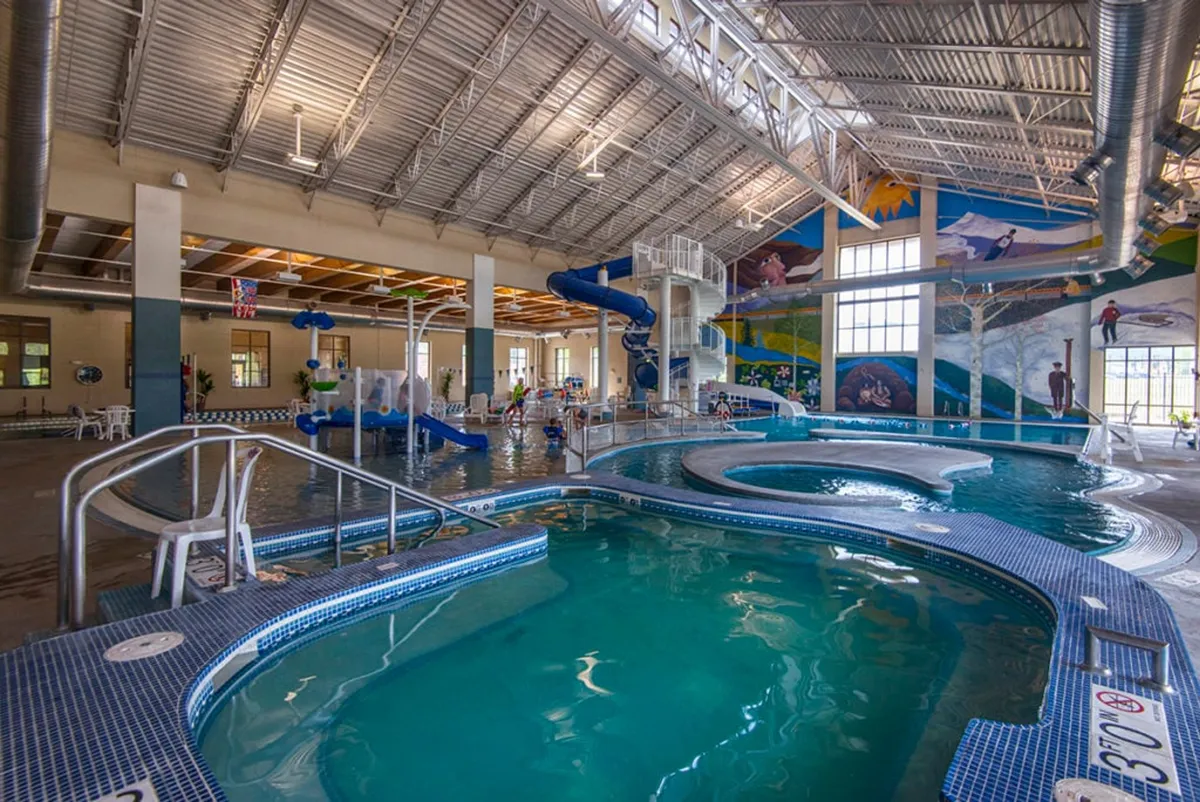 A vibrant indoor pool area featuring a central pool, colorful wall murals, water slides, children actively playing, and lounge chairs where parents and families can relax. The playful atmosphere and family-oriented amenities are clear, making it an inviting space for children and adults alike.