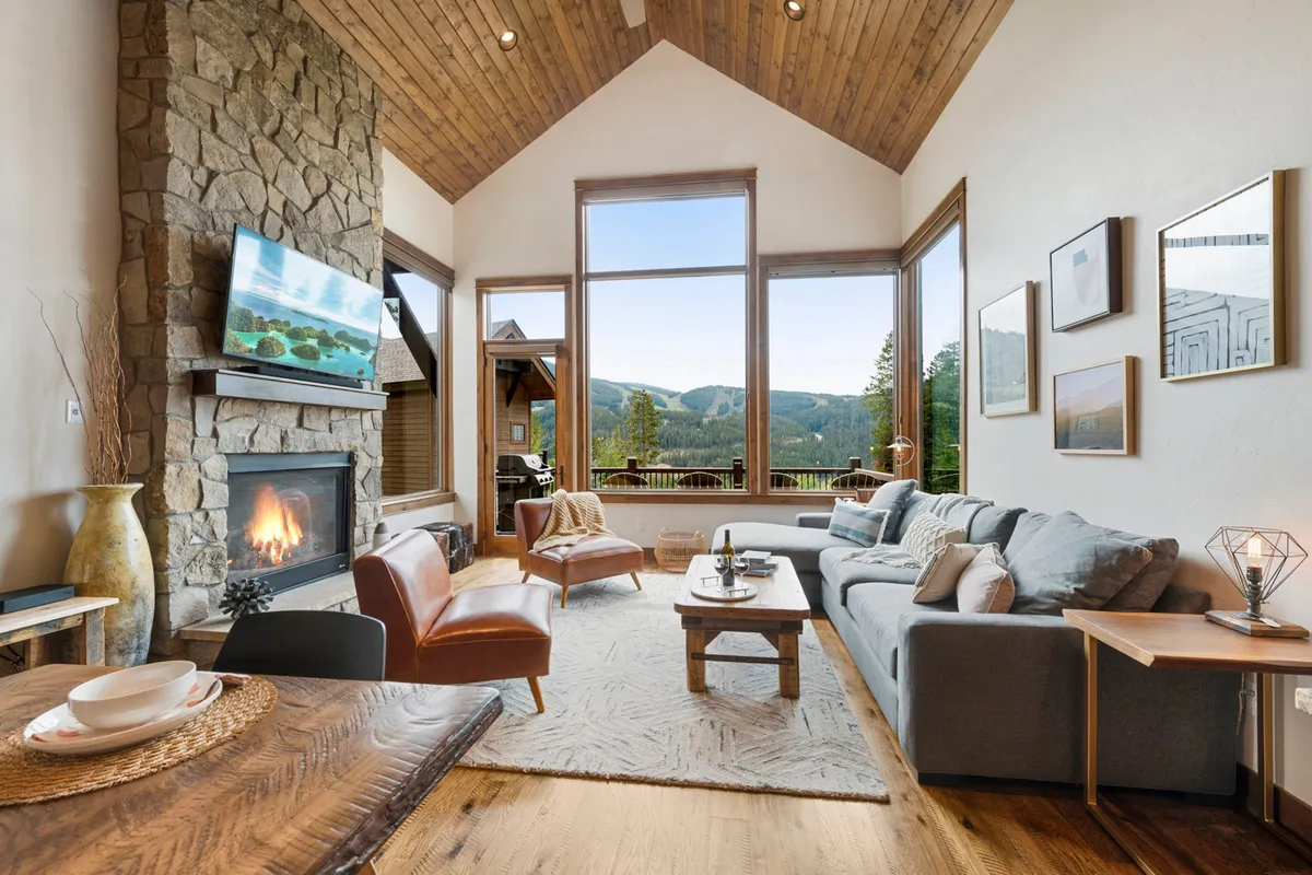 A spacious living room featuring a striking stone fireplace, large windows that let in abundant natural light, comfortable seating areas, and a wooden coffee table. Through the windows, breathtaking mountain views are visible, merging the indoors with the scenic outdoors.
