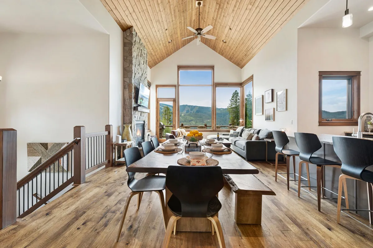 A bright, open dining and kitchen area with large windows framing mountain vistas, a dining table set for a family meal, modern appliances, and a wooden ceiling with exposed beams. The seamless transition between kitchen, dining, and living spaces is clear.
